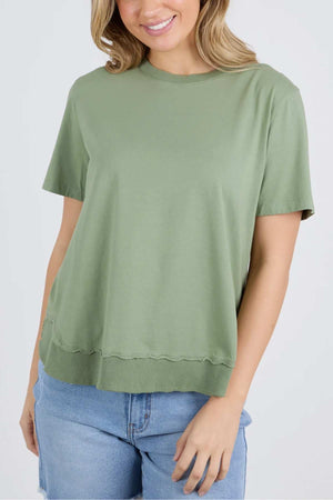 A person is wearing a casual outfit consisting of the Foxwood Farrah Short Sleeve Tee in light green and blue denim shorts. The t-shirt has a relaxed fit with a slightly wavy hem. The person has shoulder-length, light brown hair and is smiling softly. The background is plain white.