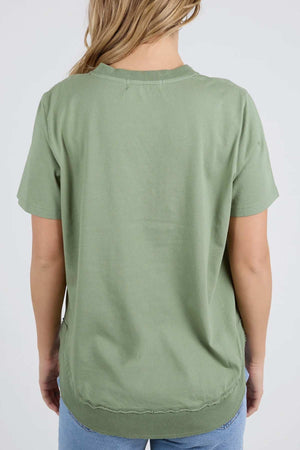 A person with long blonde hair is seen from the back, wearing a Foxwood Farrah Short Sleeve Tee in light green and light blue jeans.