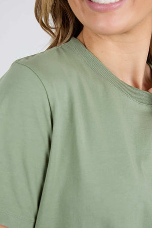 A person with light skin is partially visible from the shoulders up, smiling while wearing a Foxwood Farrah Short Sleeve Tee in light green. The background is plain and out of focus.