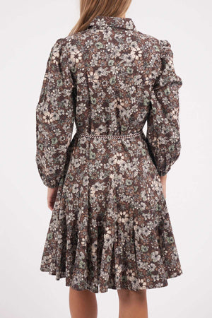 A woman with straight, light brown hair is seen from the back wearing a knee-length Foxwood Floral Meadow Dress featuring a brown floral print, long sleeves, a fitted drawstring waist, and a flared skirt.