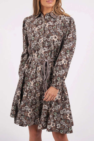 Wearing the Foxwood Floral Meadow Dress, featuring long sleeves and a brown floral print with a drawstring waist and flared skirt, a woman stands with her hands by her sides, face partially out of frame.