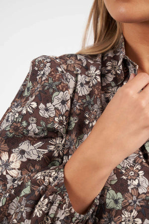 A close-up shows someone adjusting the collar of a brown, green, and white floral print shirt, revealing only part of their face and upper body. The look is inspired by the Foxwood Floral Meadow Dress.