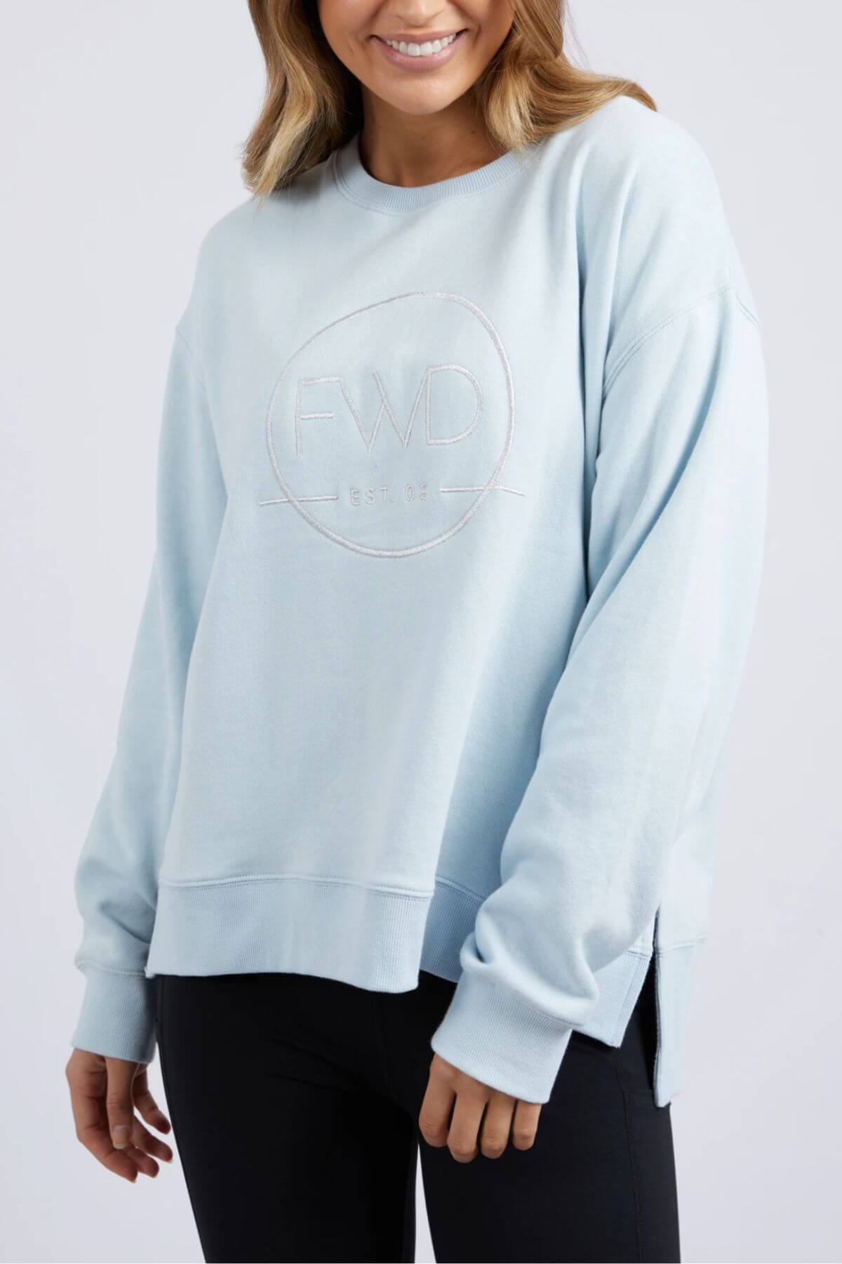 A woman with light brown hair, wearing black pants and smiling, is dressed in a light blue Foxwood Formation Crew sweatshirt. The sweatshirt, made from 100% cotton and offering a relaxed fit, features the letters "FWD" and "EST. 07" embossed on the front.