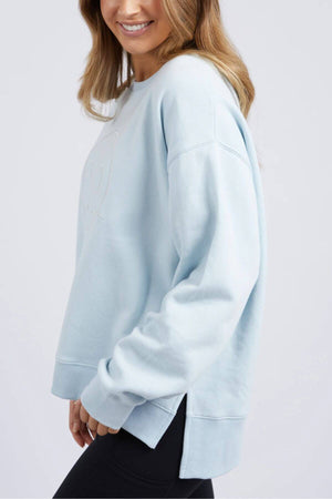 A person, shown from the shoulders down, is standing against a plain background wearing a light blue Foxwood Formation Crew sweatshirt and black leggings. The 100% cotton sweatshirt, designed for a casual look, features slightly dropped shoulders and a side slit hem in a relaxed fit.