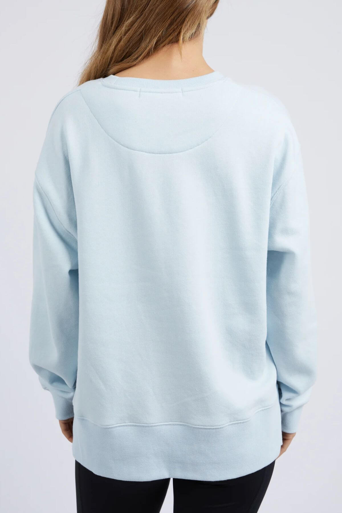 A person with long hair is seen from behind wearing a light blue Foxwood Formation Crew sweatshirt made of 100% cotton. The relaxed fit of the sweatshirt showcases a simple design with a round neckline and long sleeves. They are also wearing black pants, set against a plain and neutral background.