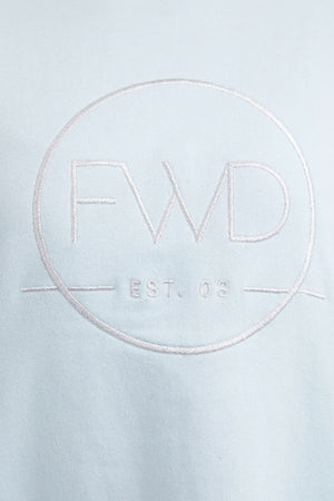 Close-up of a light blue fabric from the Foxwood Formation Crew showcasing an embroidered circular logo. The logo prominently displays the letters "FWD" in large font, with "EST. 03" written beneath it, separated by a horizontal line. The stitching is in a slightly darker shade than the 100% cotton fabric, contributing to its relaxed fit aesthetic.