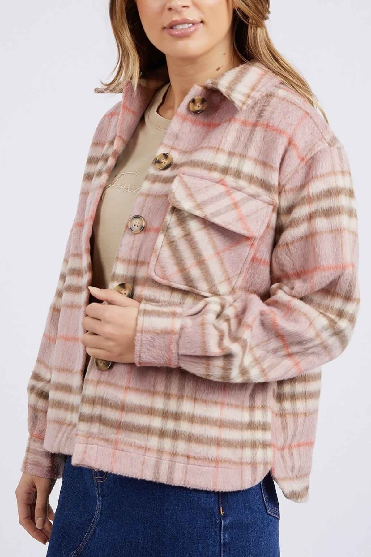 A person is dressed in a pink and beige plaid shacket similar to the Foxwood Frankie Check Coat, featuring large buttons and a chest pocket. Its soft texture goes well with their light brown t-shirt and blue jeans, while the yarn-dyed check pattern contrasts with the plain white background.