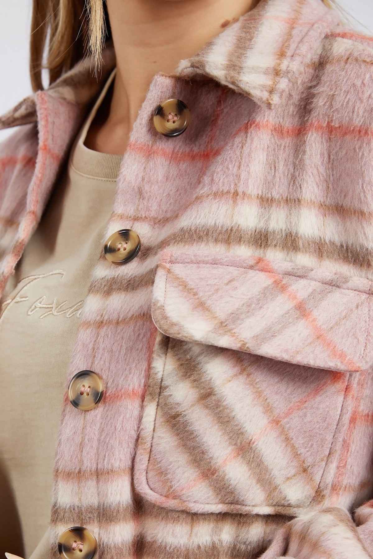 Close-up of a person wearing the Foxwood Frankie Check Coat, showcasing its yarn-dyed pink, white, and tan check design with large buttons and a chest pocket. Underneath is a beige T-shirt with subtle writing. Only part of the person's face and neck are visible.