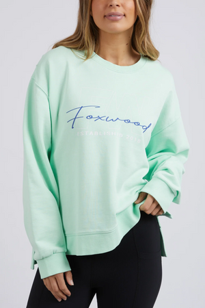 Someone is wearing a Foxwood Glider Crew sweatshirt in light mint green, with "Foxwood" in blue cursive. It's a relaxed-fit piece made of 100% cotton, paired with black pants against a plain, light-colored background.