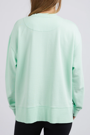 Someone with long hair wears an oversized Foxwood Glider Crew in light green, showcasing a relaxed fit made from 100% cotton. They're paired with black pants and face away against a plain white background.
