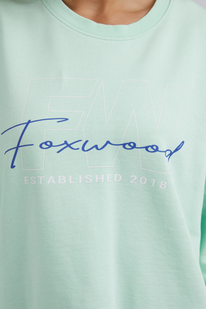 The Foxwood Glider Crew showcases a light green design with "Foxwood" in blue cursive over faint "FW" letters. Below, "ESTABLISHED 2018" appears in white. Made from 100% cotton, this relaxed-fit shirt offers ultimate comfort and style.