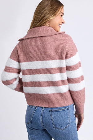 A woman with long blonde hair wears the Foxwood Gracie Zip Crew, a pink sweater with white stripes, paired with blue jeans. She faces away, looking over her shoulder and smiling against a plain white background.