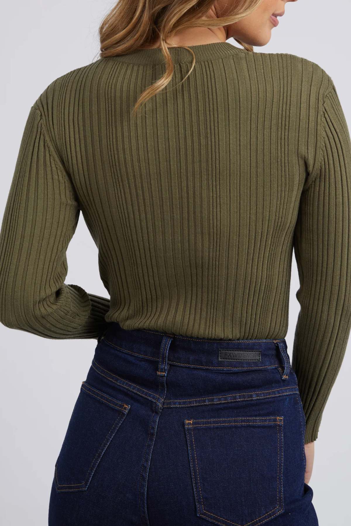 Standing against a light gray background, a person with light brown wavy hair wears dark blue jeans and a Foxwood Greta Long Sleeve sweater in ribbed olive green, offering both comfort and durability.