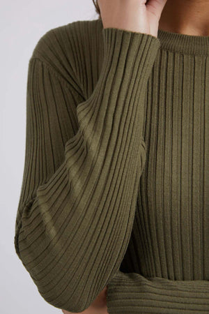 An up-close image shows someone in the Foxwood Greta Long Sleeve sweater in ribbed olive-green, their hand gently touching their face. The soft ribbed fabric emphasizes comfort and durability, featuring textured details and cozy long sleeves against a simple light background.