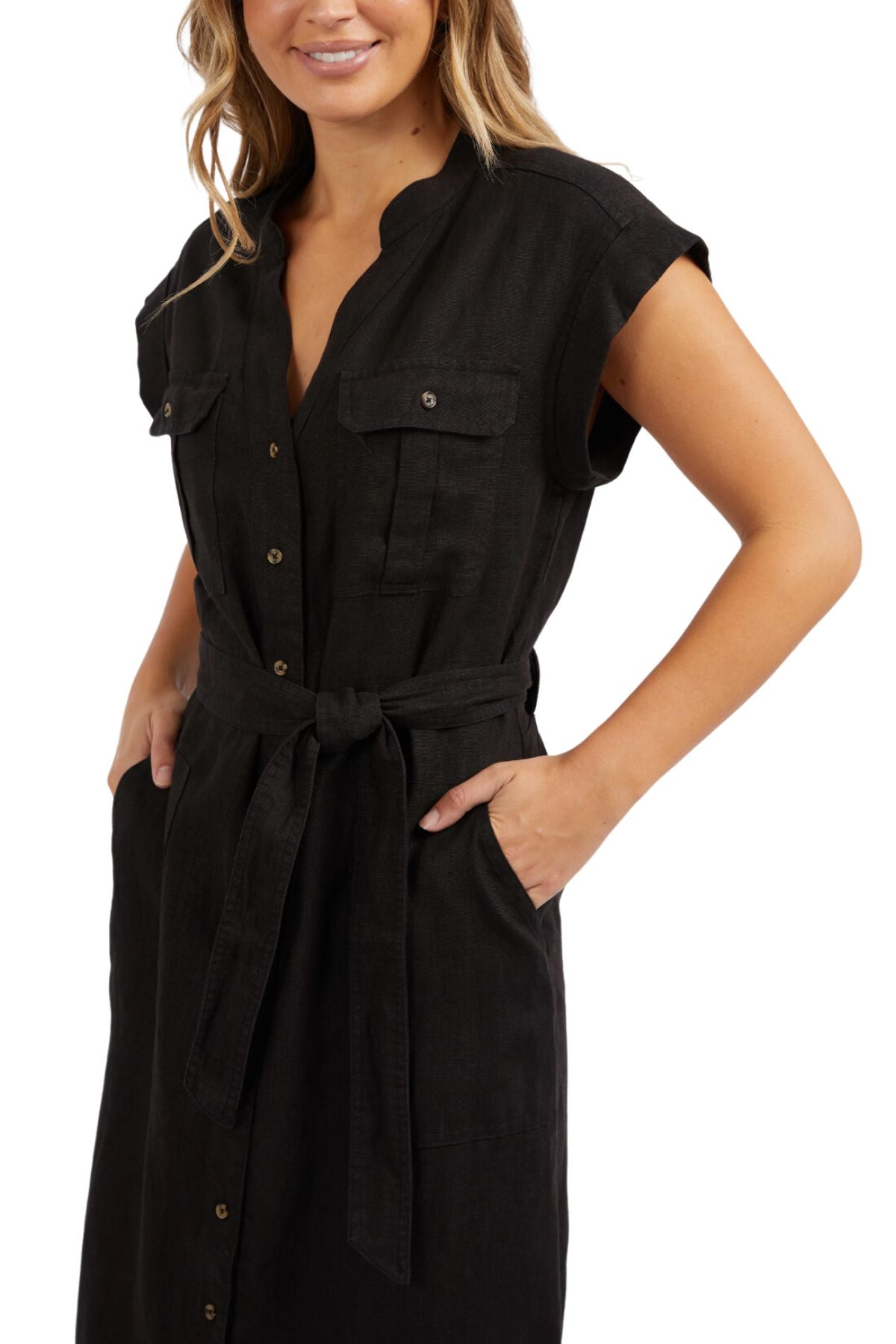 The woman is wearing the Foxwood Harlow Midi Dress, a black linen dress in a shirt-style with a button-up front, V-neck, and short sleeves. It features a waist-cinching belt and two chest pockets. She smiles warmly, her hands resting casually in the dress pockets.