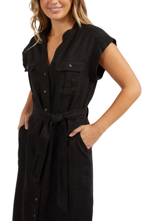 The woman is wearing the Foxwood Harlow Midi Dress, a black linen dress in a shirt-style with a button-up front, V-neck, and short sleeves. It features a waist-cinching belt and two chest pockets. She smiles warmly, her hands resting casually in the dress pockets.