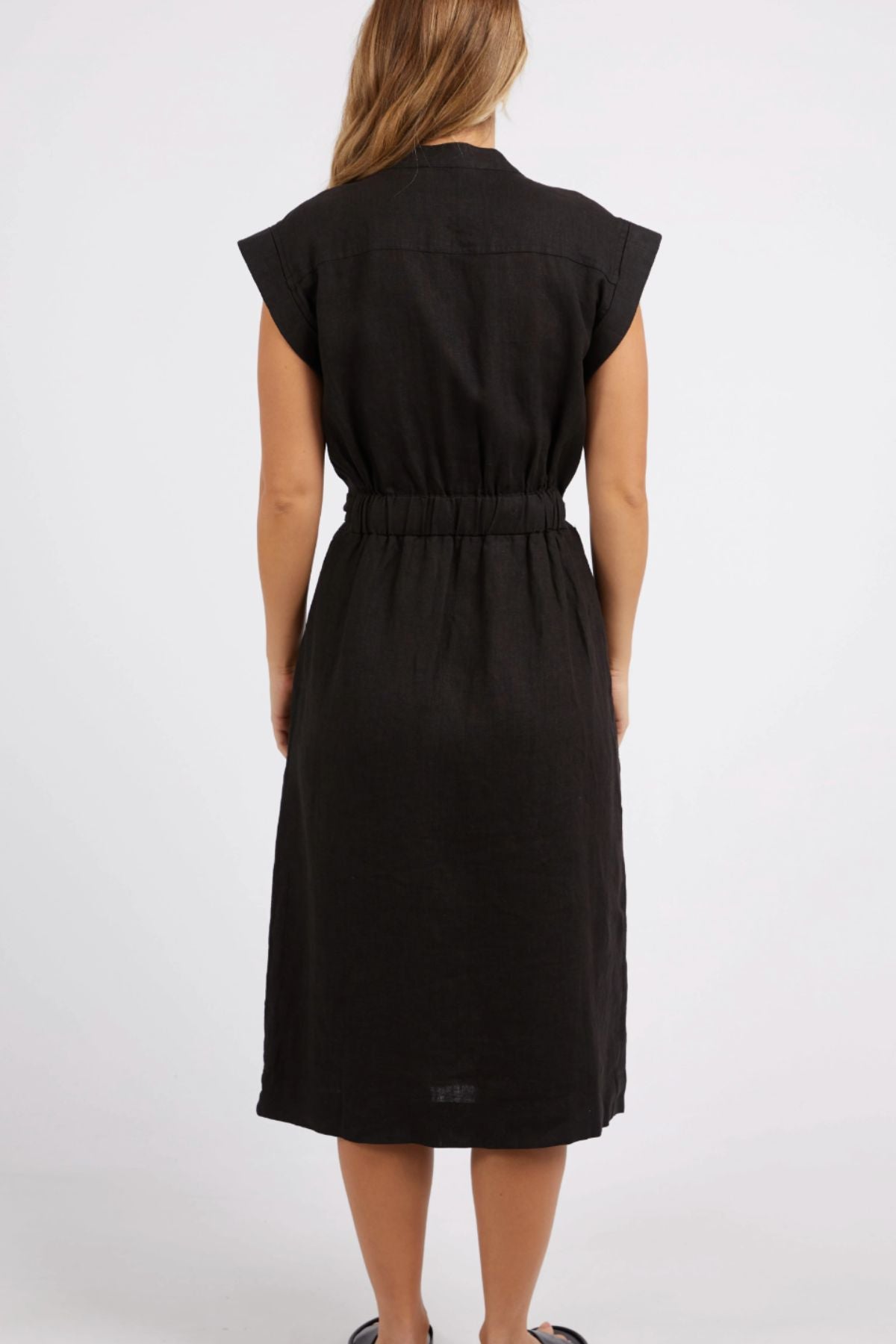 A woman with long hair is wearing a black, sleeveless Foxwood Harlow Midi Dress featuring a gathered waist. She stands with her back to the camera against a plain white background.
