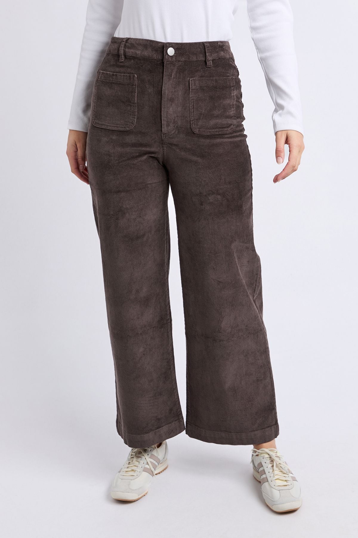Model wears the Foxwood Henrietta Cord Pant in brown (high-rise, wide-leg, large front pockets) with a white long-sleeve shirt and white sneakers, posed against a plain light background.