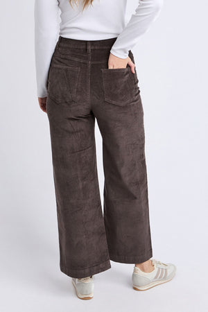 Wearing a long-sleeve white top, Foxwood Henrietta Cord Pant in brown, and white sneakers, a person stands facing away with one hand in their back pocket against a plain background.