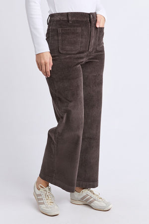 A person wearing the Foxwood Henrietta Cord Pant in brown, a white long-sleeve shirt, and white sneakers with gray stripes stands against a plain white background.