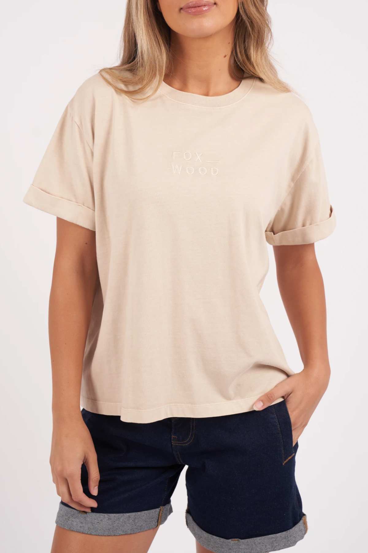 A person is wearing a Foxwood Huntleigh Oversized Tee in beige with rolled-up sleeves and "FOX WOOD" printed on the chest. They are also sporting denim shorts with rolled-up hems, have one hand in their pocket, and are standing against a plain background.