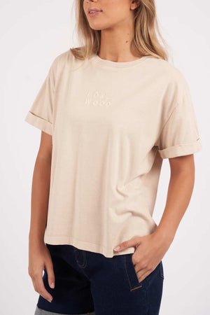 A person with long blonde hair is wearing a beige Foxwood Huntleigh Oversized Tee that reads "FOX WOOD" in small, subtle text on the front. They are also dressed in dark blue jeans and have their right hand in their pocket, standing against a plain white background.