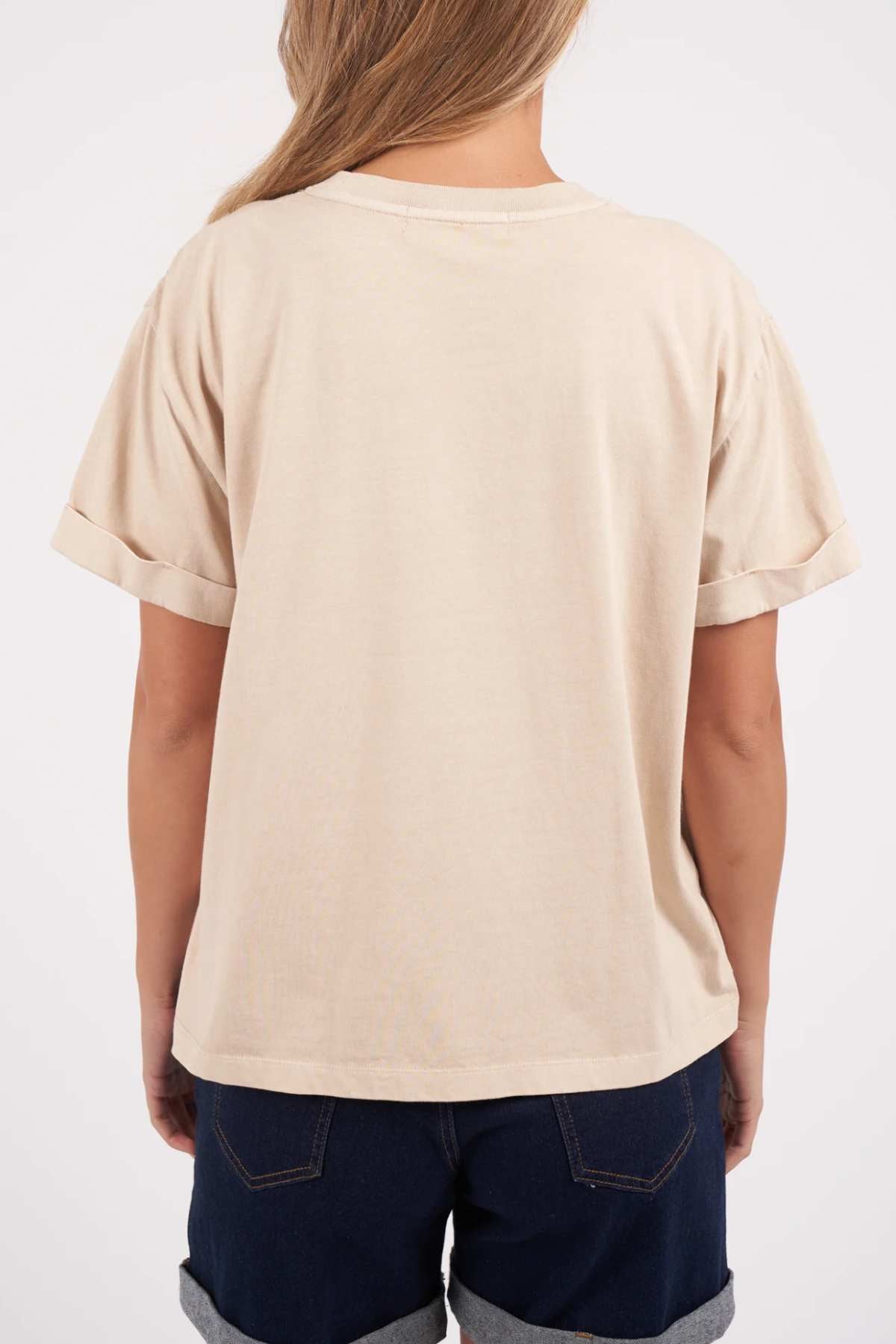 A person with long hair is seen from the back, wearing a Foxwood Huntleigh Oversized Tee in beige and dark denim shorts. The t-shirt has rolled-up sleeves, and the person's hair falls naturally over their shoulders. The background is plain and light-colored.