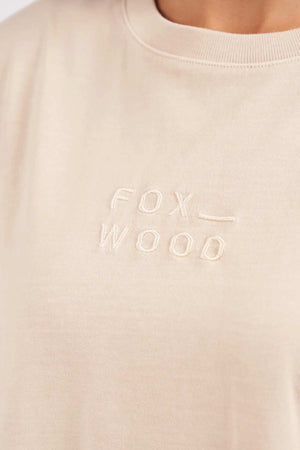 Close-up image of the Foxwood Huntleigh Oversized Tee in beige, featuring the word "FOXWOOD" embroidered in slightly darker beige thread on the chest. Only a portion of the person's neck and shoulders are visible.