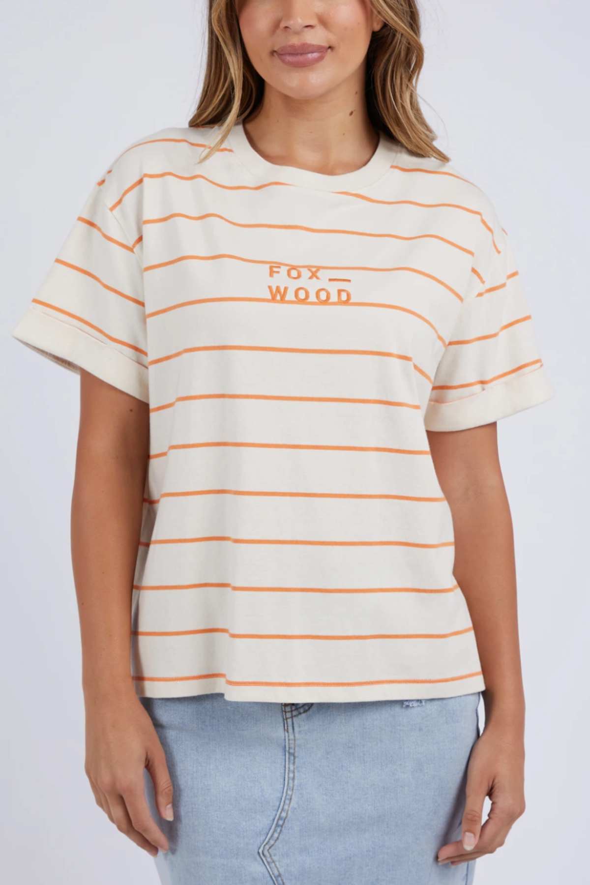 A person wearing a beige Foxwood Huntleigh Stripe Tee, featuring orange horizontal stripes and "Fox Wood" text on the front, stands against a plain background. The 100% cotton T-shirt boasts a relaxed fit, and the individual pairs it with a light blue denim skirt. Only the torso and partial arms are visible.