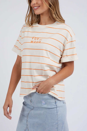 The individual is wearing a Foxwood Huntleigh Stripe Tee, an oversized T-shirt made from 100% cotton with orange horizontal stripes and "FOX WOOD" emblazoned across the front. The shirt is tucked into a light blue, distressed denim skirt. They have a smile on their face and one hand in their skirt pocket.