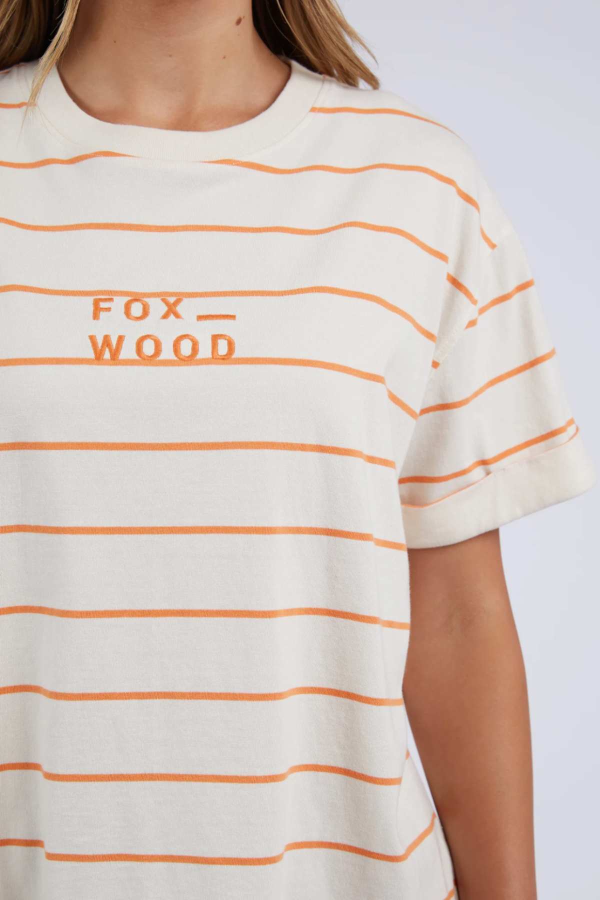 A person is wearing the Foxwood Huntleigh Stripe Tee, a cream-colored short-sleeve shirt with orange horizontal stripes and "FOX WOOD" embroidered on the front, made from 100% cotton. Only the torso and part of the arm are visible against a plain background.
