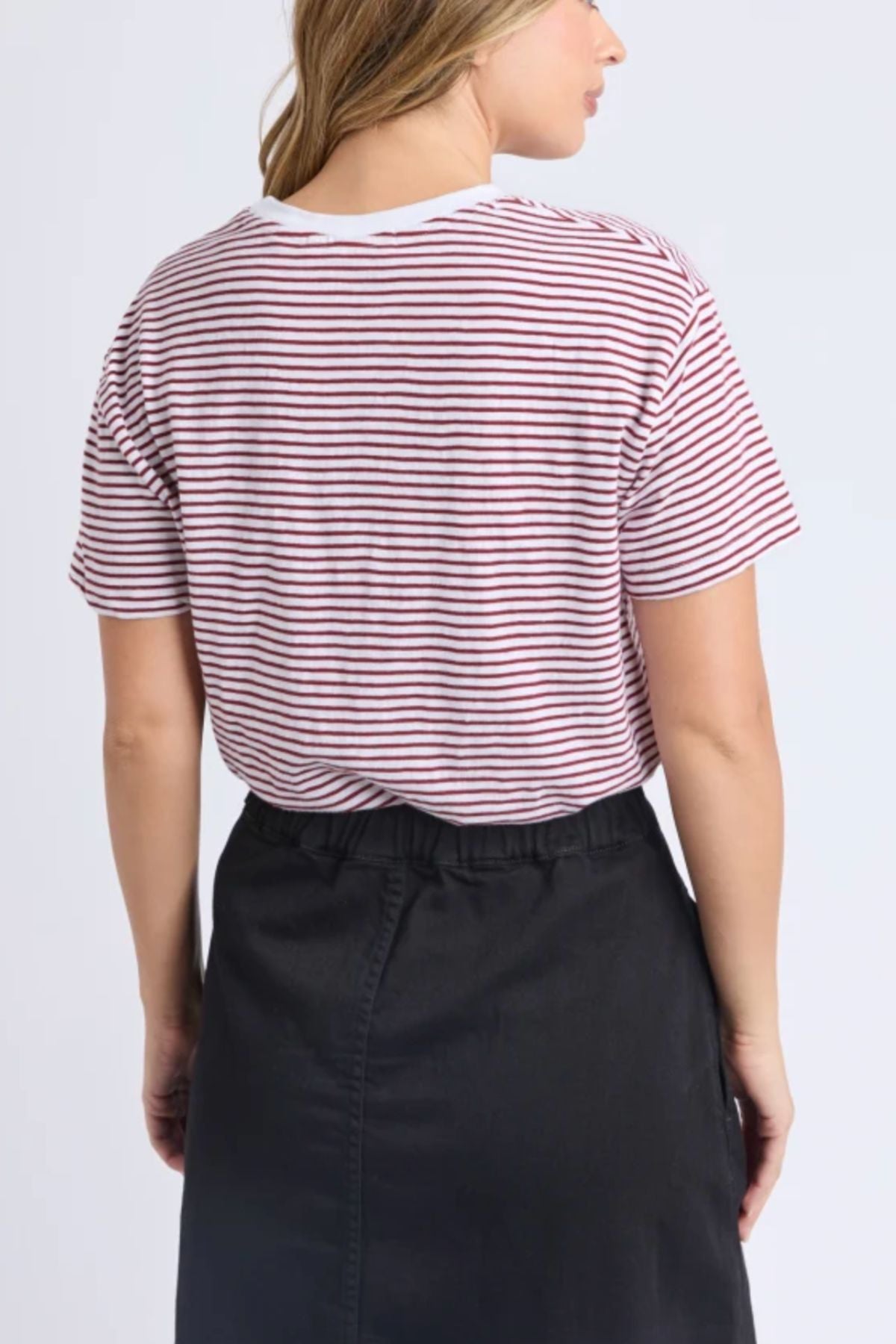 A woman faces away from the camera in the Foxwood Hutch Stripe Tee, featuring thin red stripes and a relaxed fit, paired with a high-waisted black skirt. Her light brown hair completes an effortlessly stylish look for daily wear.