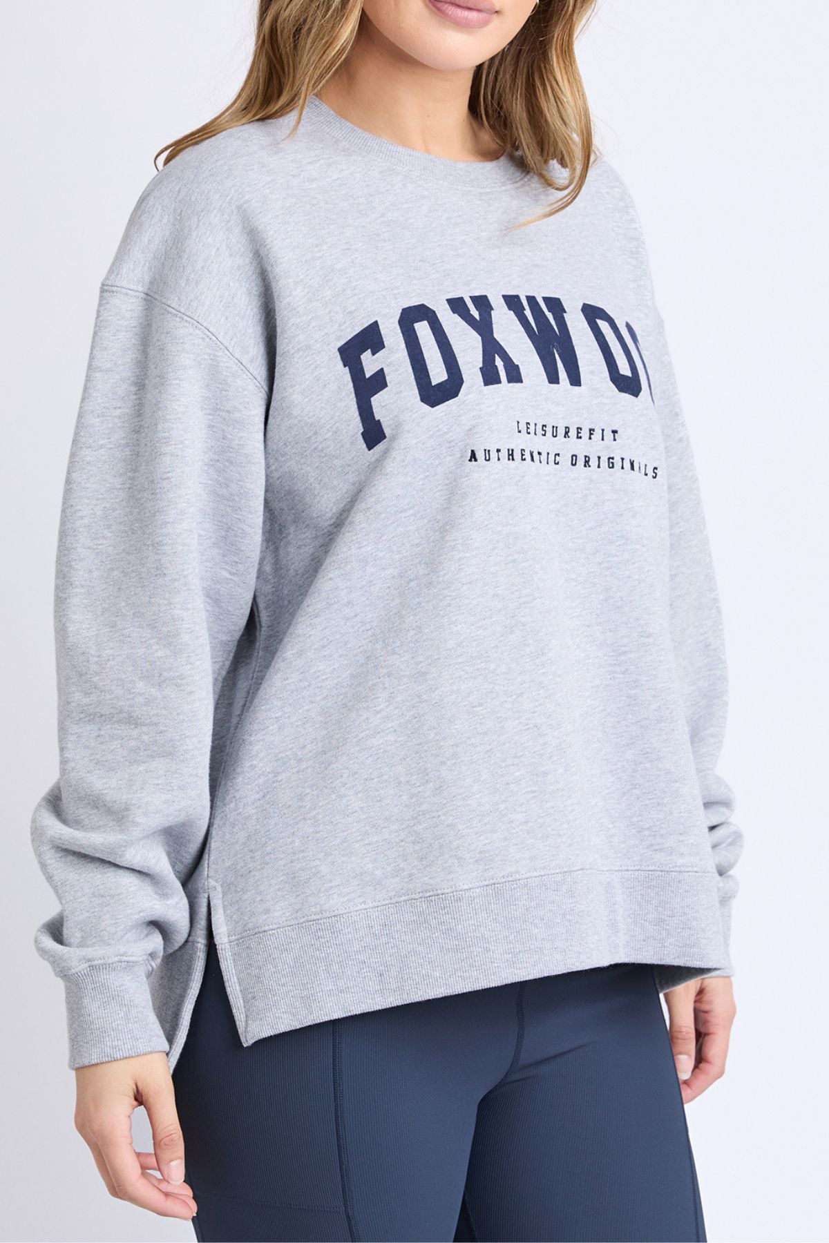 Wearing the Foxwood Intention Crew sweatshirt in light gray cotton-rich fleece with bold navy "FOXWOOD" lettering, paired with dark blue leggings, someone stands against a plain light background.