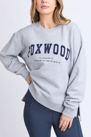 Wearing the Foxwood Intention Crewβa cotton-rich, light gray relaxed fit fleece featuring "FOXWOOD LEISUREFIT AUTHENTIC ORIGINALS" in navyβa person pairs it with dark gray leggings and poses with one hand on their hip.