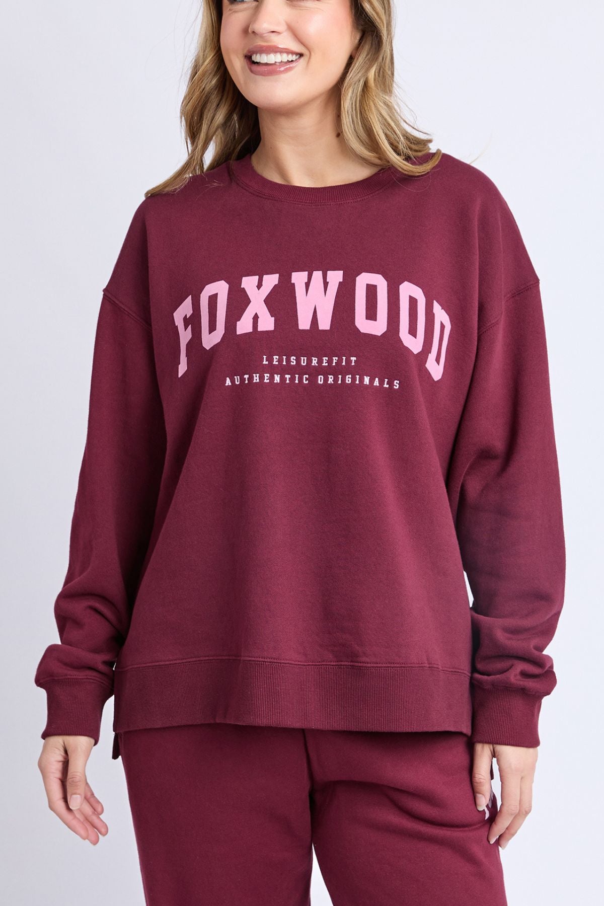 A smiling person wears the maroon Foxwood Intention Crew and matching pants. The relaxed-fit cotton-rich fleece sweatshirt features "FOXWOOD" in large pink letters and "LEISUREFIT AUTHENTIC ORIGINALS" below.