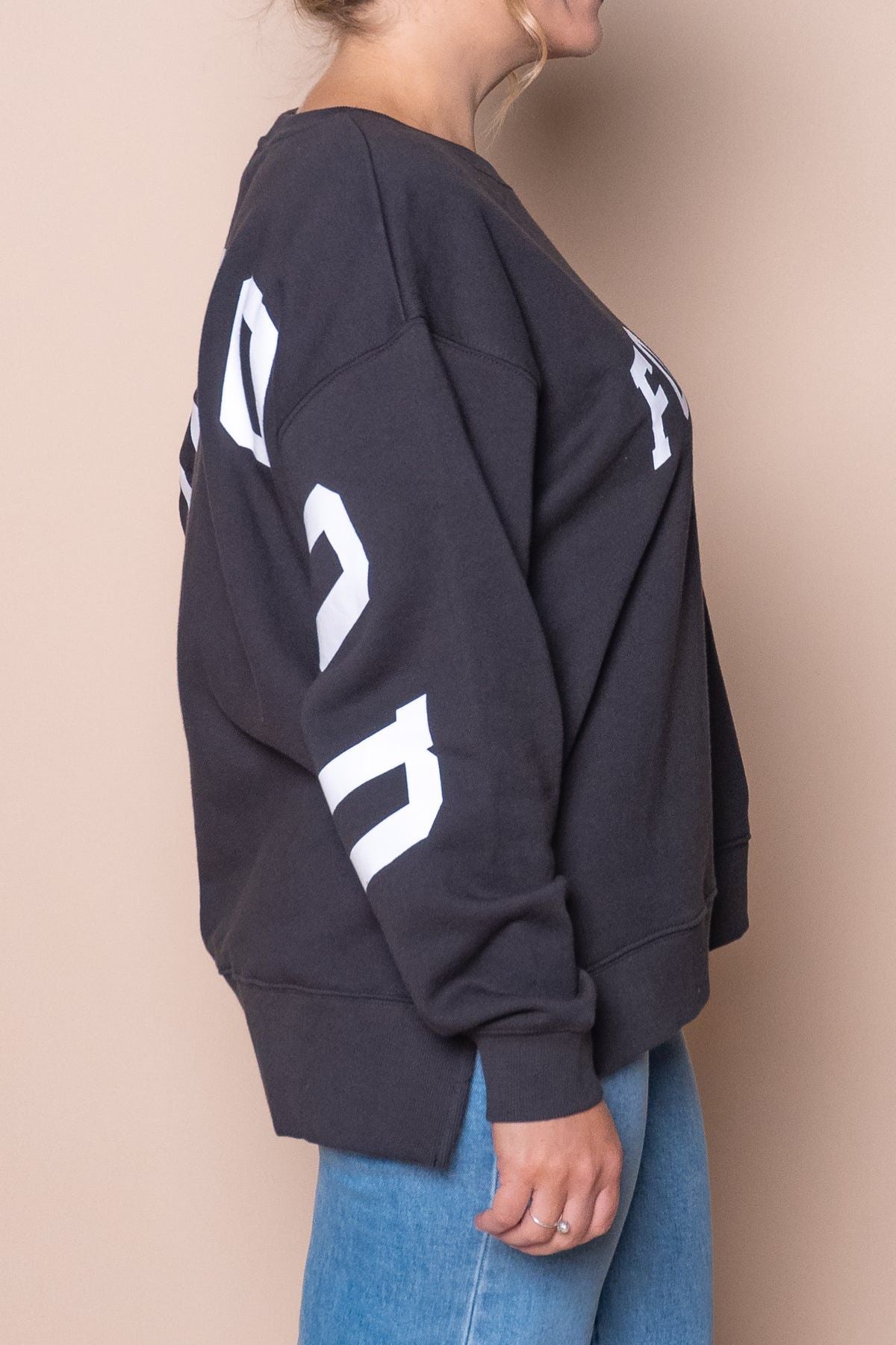 A person in a relaxed-fit Foxwood Interval Crew fleece in black with white lettering on the chest and sleeve, paired with light blue jeans, stands sideways against a beige background—an everyday wardrobe essential.