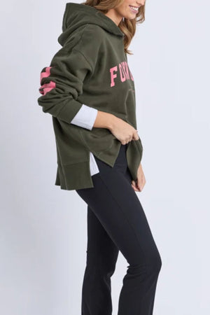 A person wearing the Foxwood Interval Hoodie with pink lettering, a white shirt underneath, and black pants stands sideways, smiling and casually holding the hem—a stylish choice for everyday wear.