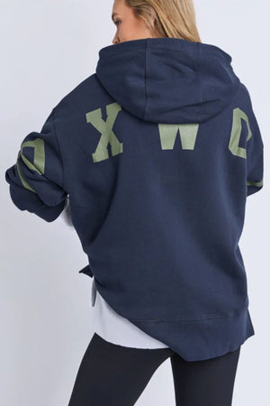 A person with long blonde hair, facing away from the camera, wears the Foxwood Interval Hoodie in a relaxed fit with large olive green "XWC" logo on the back, paired with black leggings and a white shirt showing underneath.