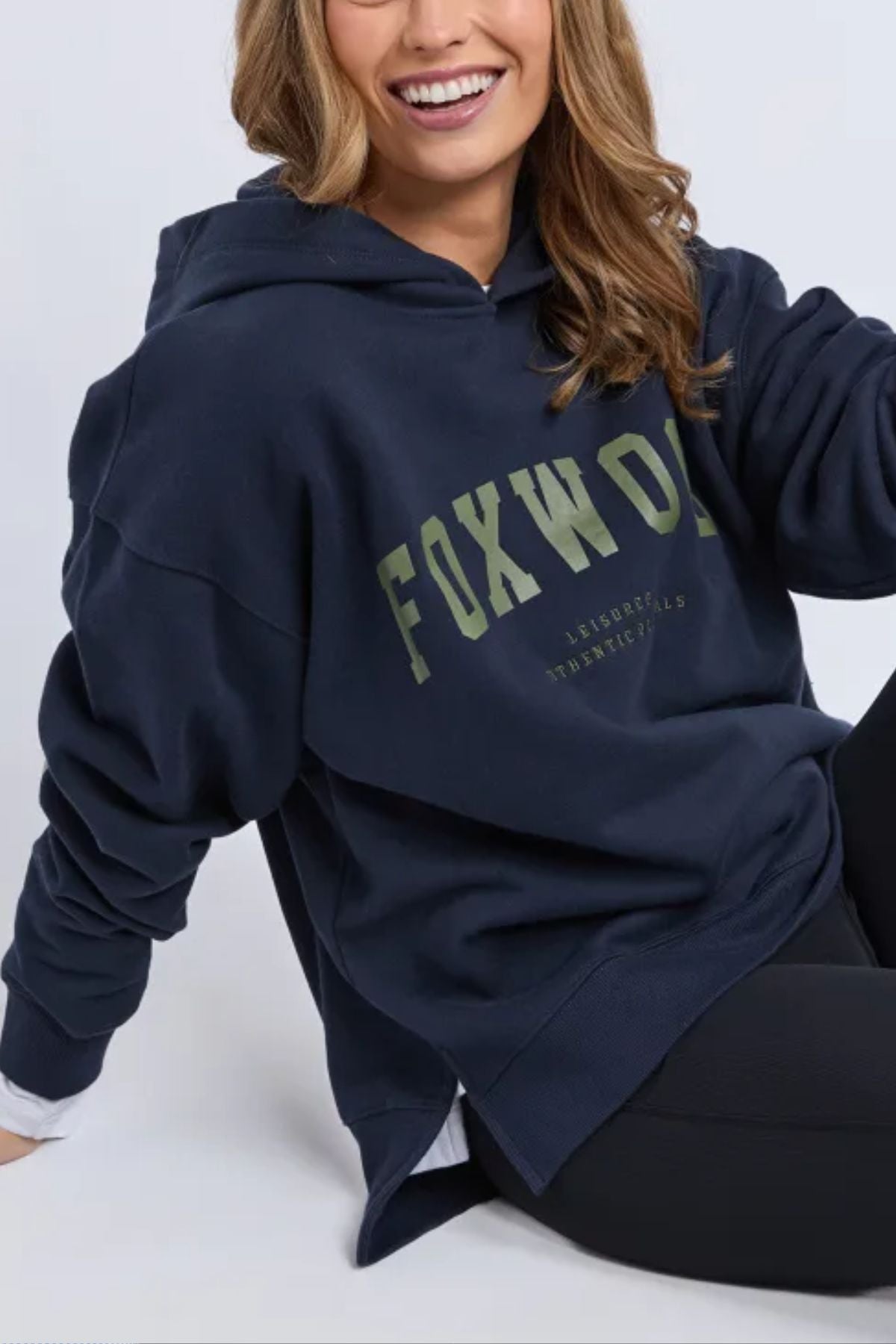 A woman with wavy hair smiles while seated, wearing a relaxed navy Foxwood Interval Hoodie featuring front logo branding, layered over a white shirt and paired with black leggings.