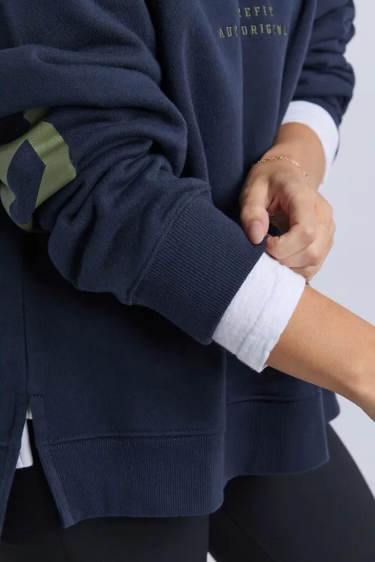 Wearing a Foxwood Interval Hoodie, an individual rolls up their sleeve to reveal part of their forearm over a white long-sleeve shirt. The focus is on the torso and arms, showcasing the hoodie's relaxed fit.