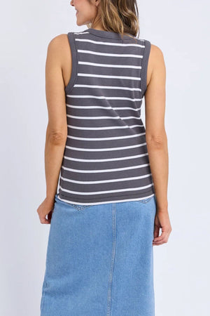 A woman with loose light brown hair faces away, wearing a relaxed fit gray and white Foxwood Iris Stripe Tank and a light blue denim skirt. Her arms rest at her sides against a plain background.