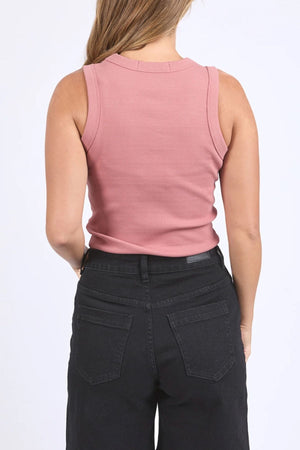 A woman with loose brown hair is shown from the back wearing the Foxwood Iris Tank, a sleeveless, ribbed stretch pink cotton top, paired with high-waisted black jeans against a plain white background.
