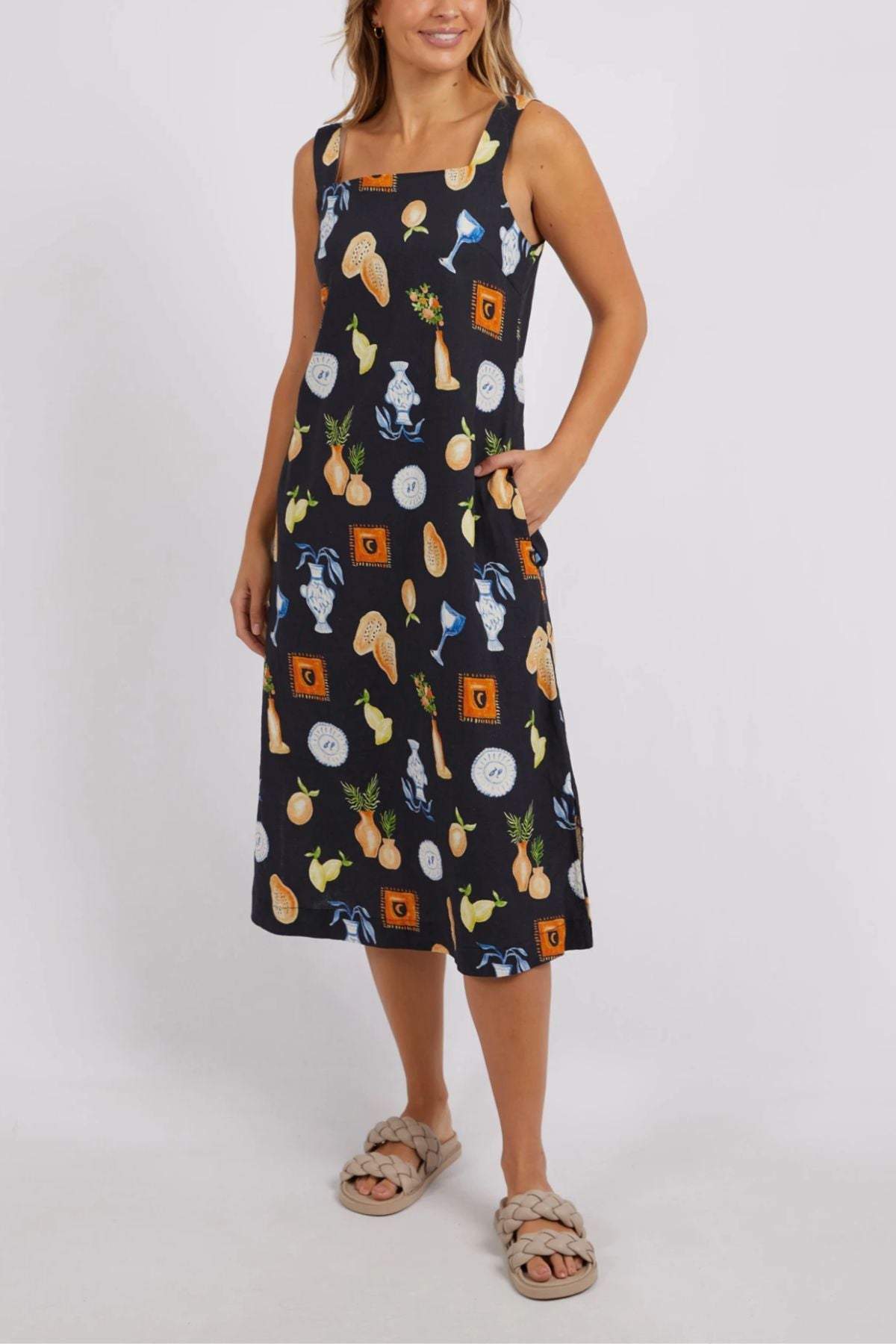 A person stands against a plain background wearing the Foxwood Italian Summer Dress, featuring a vibrant pattern of cats, vegetables, and bread. Their stylish summer party dress is complemented with beige woven sandals.