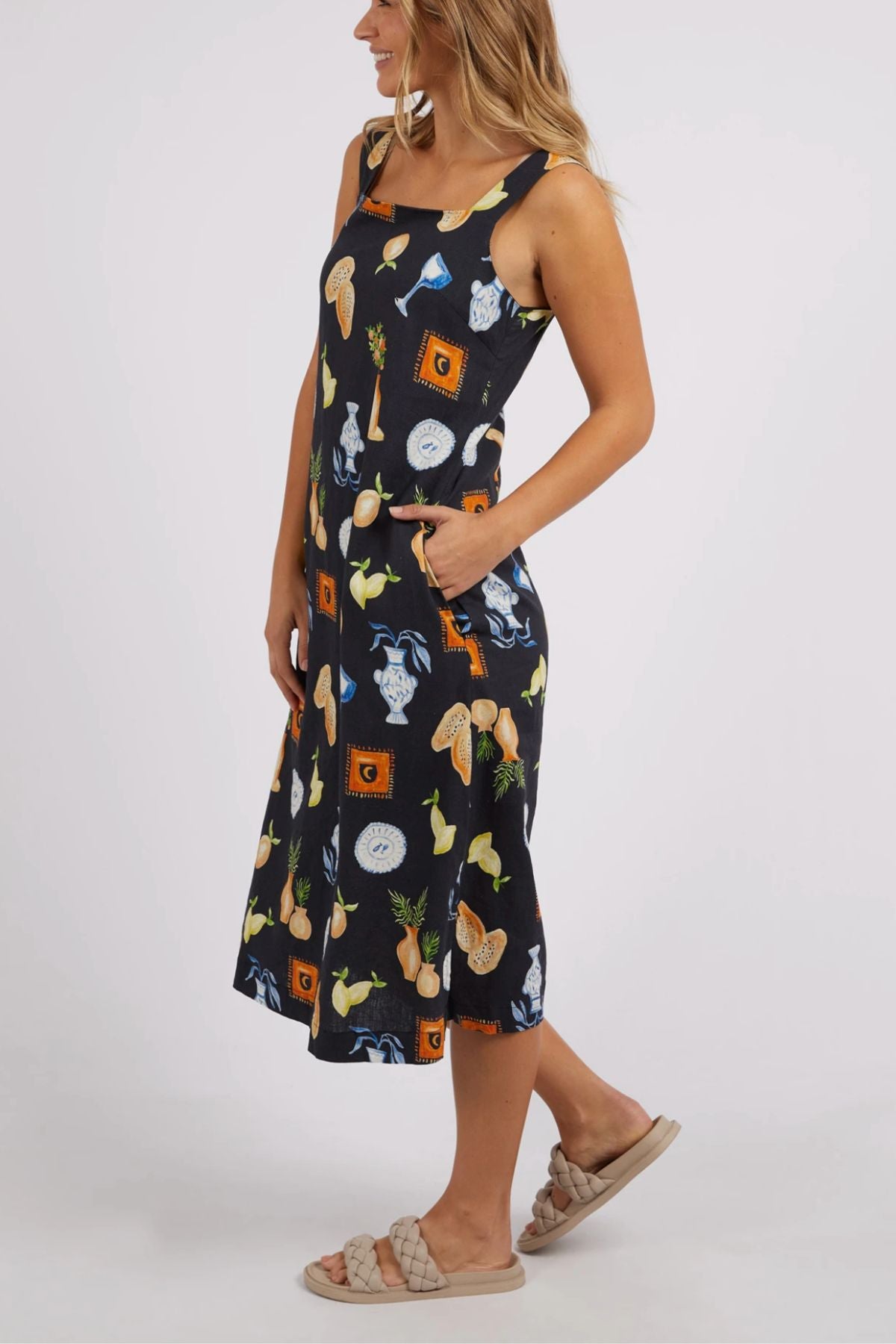 A person is dressed in a sleeveless Foxwood Italian Summer Dress in black, beautifully decorated with a lively pattern of fruits and vegetables, including pineapples and citrus. They are pictured with their hands tucked into the dress's pockets, paired with light-colored sandals—ideal for any summer celebration.