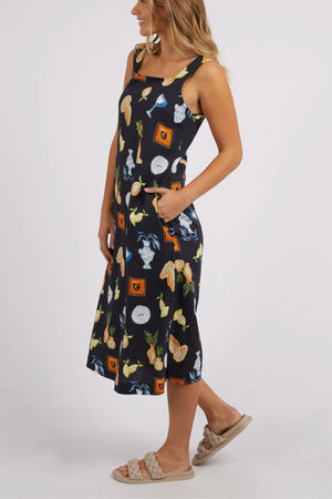 A person is dressed in a sleeveless Foxwood Italian Summer Dress in black, beautifully decorated with a lively pattern of fruits and vegetables, including pineapples and citrus. They are pictured with their hands tucked into the dress's pockets, paired with light-colored sandals—ideal for any summer celebration.