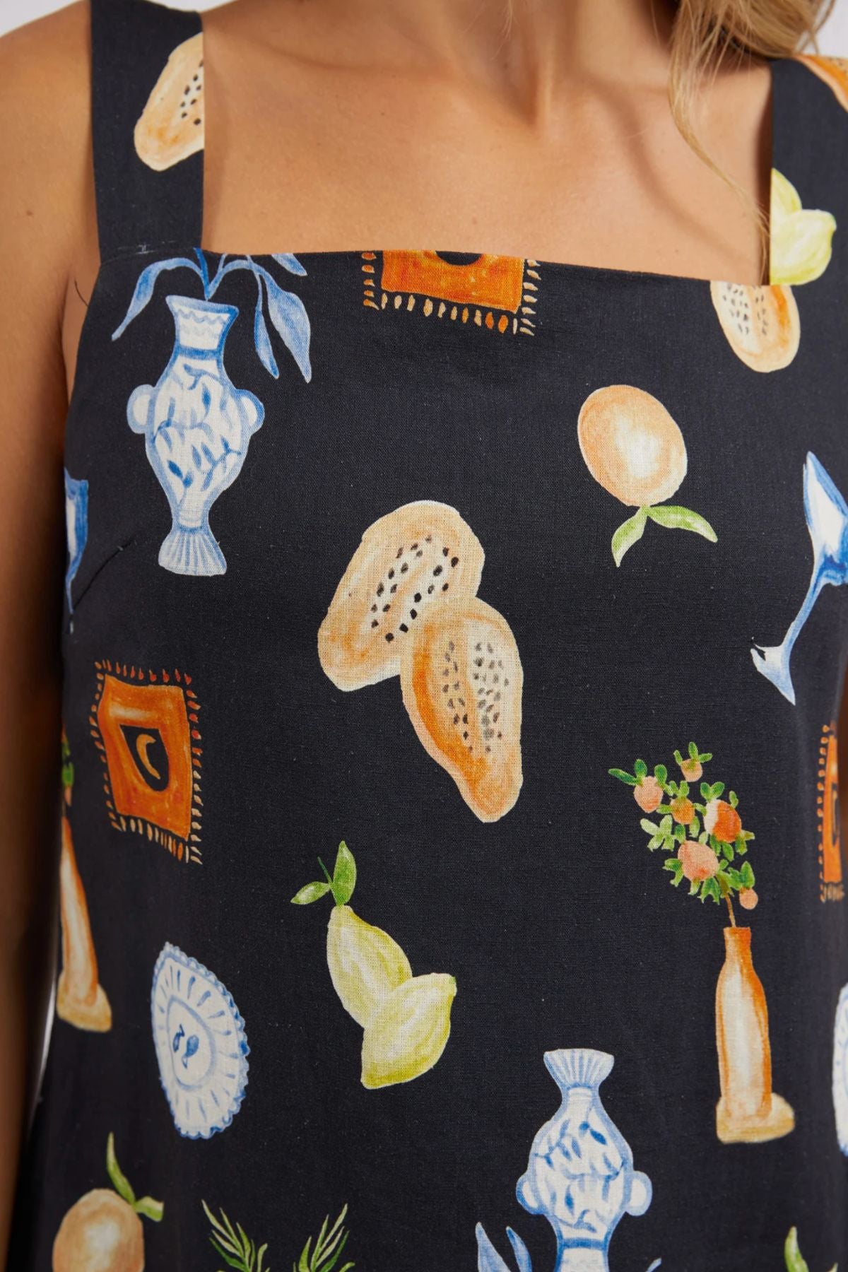 A detailed view of someone wearing the Foxwood Italian Summer Dress highlights its vibrant pattern, featuring vases, fruits, and decorative items on soft linen. This dress boasts a square neckline and thick straps, making it an ideal choice for standing out at any summer event.