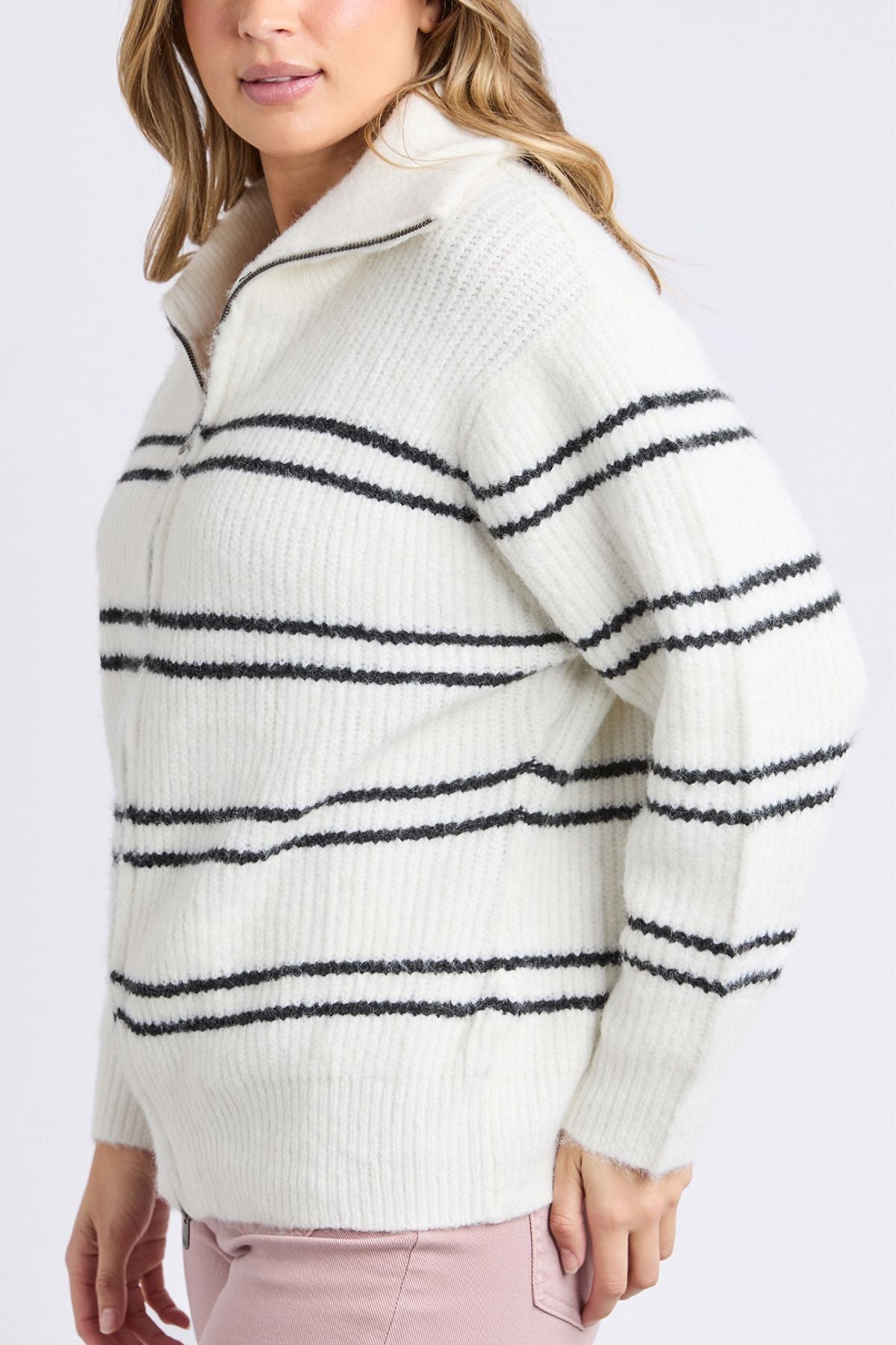 A woman wears the Foxwood Journey Zip Knit, a relaxed fit white ribbed sweater with black stripes and collar, paired with light pink pants, standing against a plain light background.