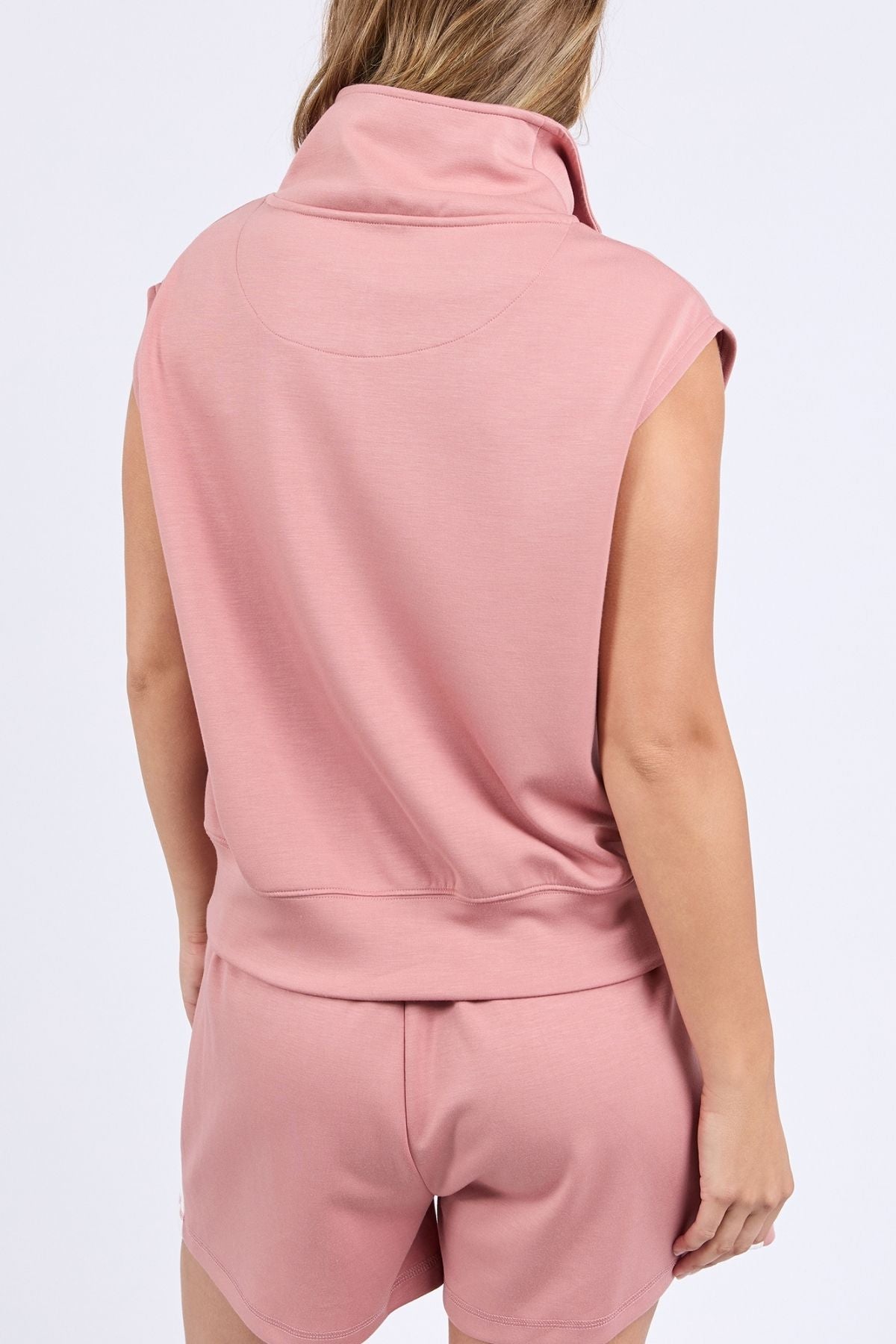 A person is seen from the back wearing a sleeveless high-collared pink top and matching shorts, styled with the Foxwood Layla Zip Vest layered on top; their left arm hangs relaxed by their side.