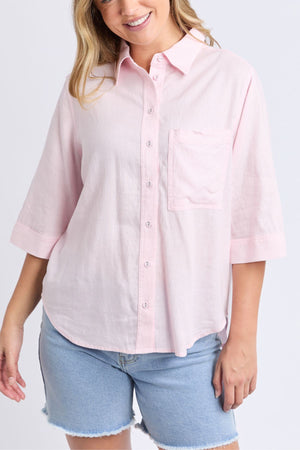 A woman wears the Foxwood Leah Shirt, a light pink linen top with a relaxed fit, chest pocket, and 3/4 sleeves, paired with light blue denim shorts, smiling against a plain background.