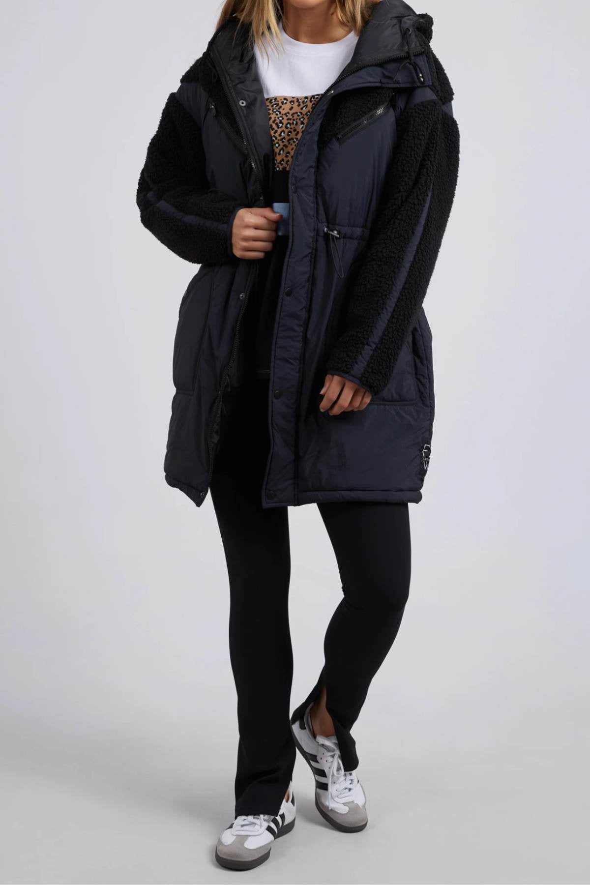 A person in a dark Foxwood Longline Anorak with faux fur sleeves, white shirt, black leggings, and black and white sneakers stands against a neutral backdrop. Made for outdoor adventure from durable nylon polyester blend, they have their hands in the coat pockets, face unseen.